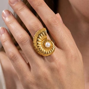 May include: A gold-toned ring featuring a textured, fan-shaped shell design with a single white pearl set at the center of the spiral. The metal surface has a polished finish with radiating grooves that mimic the natural structure of a seashell.