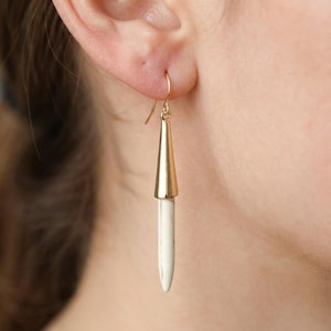 May include: A drop earring featuring a polished gold-tone metal cone cap attached to a slender, cylindrical white stone spike. The earring hangs from a simple gold-tone wire hook, creating a vertical, linear silhouette that dangles from the earlobe.