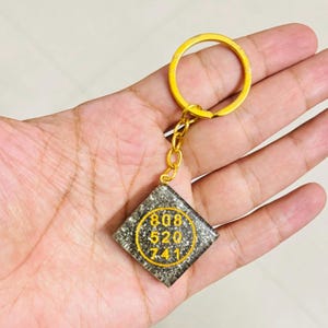 Pode incluir: Um porta-chaves quadrado com um pendente de resina escuro e brilhante, com um anel e corrente de metal dourado. O pendente apresenta uma borda circular amarela que envolve os números 808, 520 e 741 dispostos em três linhas.