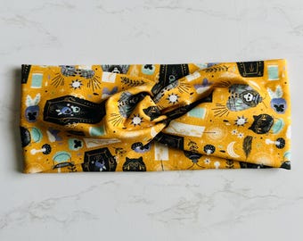 Op de afbeelding: Mosterdgele hoofdband met een herhalend patroon van zwarte doodskisten, katten, manen en paddenstoelen. De stof is aan de bovenkant geknoopt. Een comfortabel accessoire voor dagelijks gebruik.