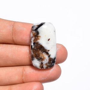 Op de afbeelding: Een witte cabochon edelsteen met bruine en zwarte insluitsels. De edelsteen is rechthoekig van vorm en heeft een glad, gepolijst oppervlak.
