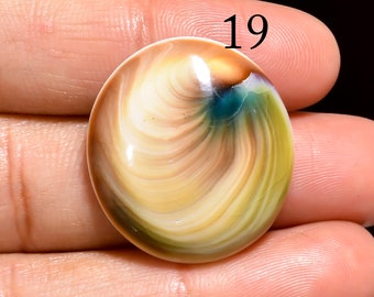 May include: A close-up of a round, multi-colored, swirling, polished stone cabochon with a light brown, white, and green pattern. The stone is being held in a hand. The number 19 is visible in the upper right corner of the image.