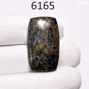 Pode incluir: Uma gema cabochão verde escura e dourada com formato retangular. A pedra tem uma superfície áspera e texturizada e está colocada sobre um fundo branco.
