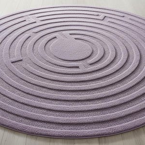 May include: A round, lavender rug with a maze-like design. The rug features concentric circles with cut-out pathways, creating a textured surface. The rug is placed on a light-colored wooden floor.