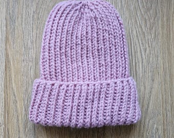 Könnte beinhalten: Eine weiche, rosafarbene Strickmütze mit Rippenstruktur. Die Mütze liegt flach auf einer Holzoberfläche.
