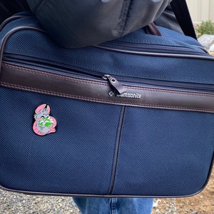 May include: A dark blue Samsonite travel bag with a brown leather trim and a zipper closure. A small pink enamel pin with a cartoon animal is attached to the bag.