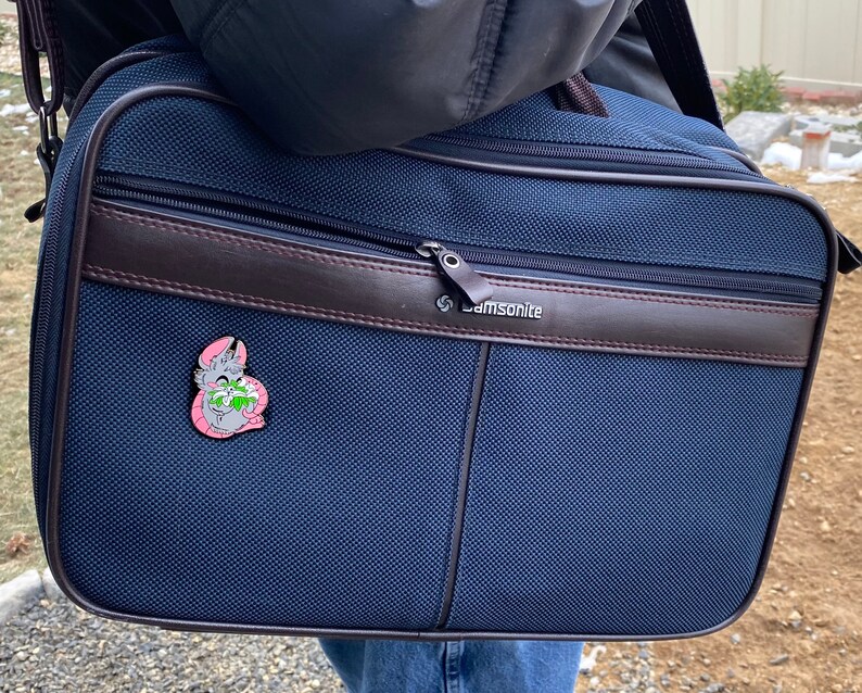 May include: A dark blue Samsonite travel bag with a brown leather trim and a zipper closure. A small pink enamel pin with a cartoon animal is attached to the bag.