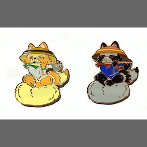 May include: Two enamel pins featuring cartoon animals playing musical instruments. The first pin is a fox wearing a straw hat and playing a flute, sitting on a yellow cloud. The second pin is a raccoon wearing a straw hat and playing a flute, sitting on a gray cloud.