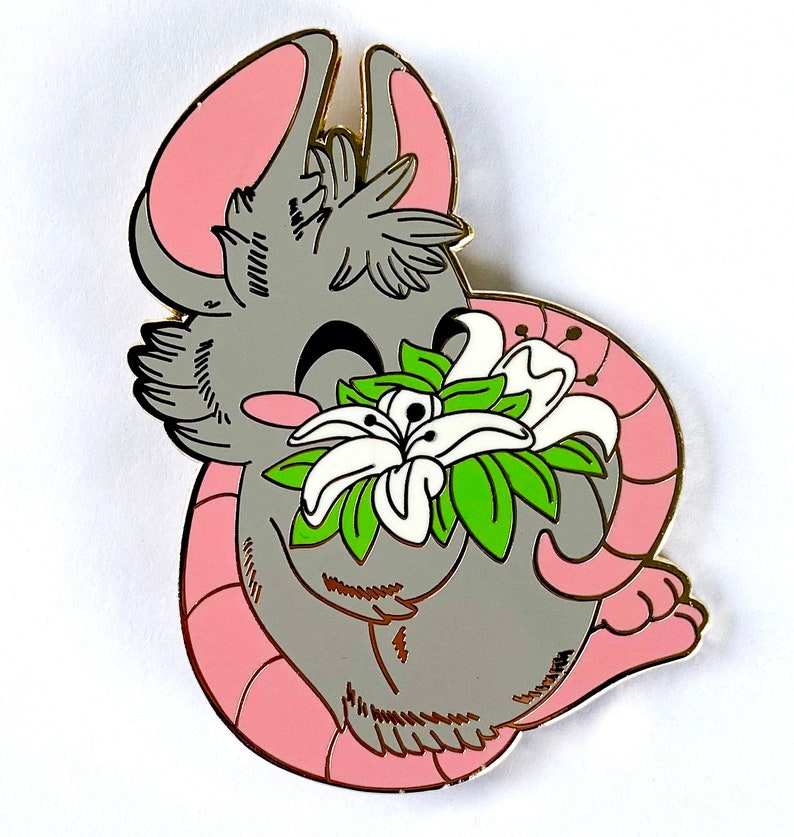 May include: An enamel pin featuring a cartoon mouse in grey and pink, holding a white flower with green leaves.