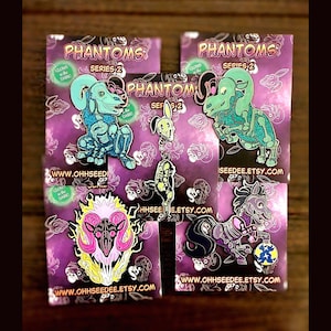 May include: A collection of enamel pins featuring the "Phantoms Series 2" designs. The pins showcase various creatures, including a dinosaur, a rabbit, a ram skull, and a horse, with skeletal and glowing details. Each pin is on a purple backing card.
