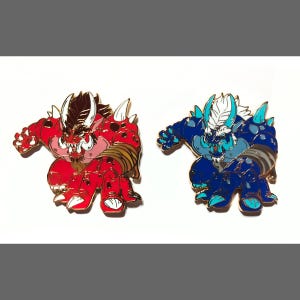 May include: Two enamel pins featuring cartoon monster characters. The first pin is red with gold accents and features a horned monster with a fierce expression. The second pin is blue with silver accents and features a horned monster with a fierce expression.