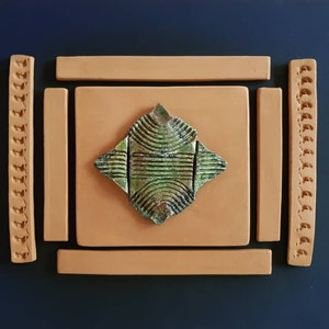 Può includere: Una piastrella di ceramica verde e marrone con un motivo geometrico è incorniciata da quattro piastrelle in terracotta. Le due piastrelle esterne hanno un bordo decorativo.
