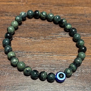 May include: A beaded bracelet featuring dark green and black speckled beads. A single blue bead with a white and black eye design is incorporated into the bracelet's design. The bracelet is displayed on a wooden surface.