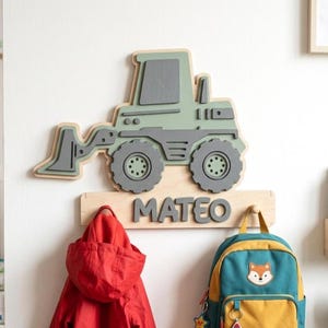 Peut inclure: Un porte-manteau mural en bois représentant un tracteur de construction superposé en vert sauge et gris foncé. La base comprend une plaque en bois naturel avec le nom MATEO en lettres capitales gris foncé en relief et deux patères.