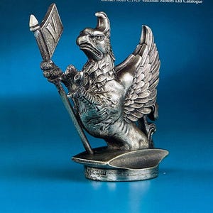 May include: A detailed silver griffin statue, holding a flag with a diamond shape. The griffin has detailed feathers and a fierce expression. The statue is mounted on a circular base against a blue background.