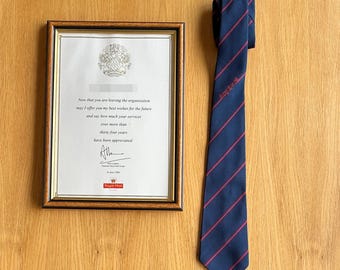 May include: A navy blue necktie with thin red diagonal stripes and the embroidered text Royal Mail, positioned next to a framed certificate featuring the Royal Mail logo, a formal message of appreciation, and a signature dated June 28, 2004.