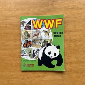 Può includere: Un album di figurine WWF con un'illustrazione di un panda su sfondo verde. La copertina mostra il logo WWF e le parole "Animali minacciati". L'album include immagini di vari animali.
