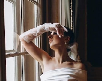 May include: A bride in a white, off-the-shoulder gown with a fitted bodice and flowing skirt. She is wearing white gloves and holding a bouquet of white roses.