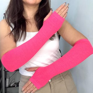 Könnte beinhalten: Ein Paar leuchtend neonpinke Armstulpen aus weichem Strickstoff. Diese langen, fingerlosen Ärmel reichen von den Knöcheln bis zum Oberarm und verfügen über eine einfache Daumenöffnung sowie eine glatte, gleichmäßige Textur.