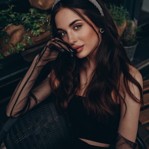 May include: A woman wearing a black top and sheer black gloves with a sparkly headband. She is sitting in a wicker chair.