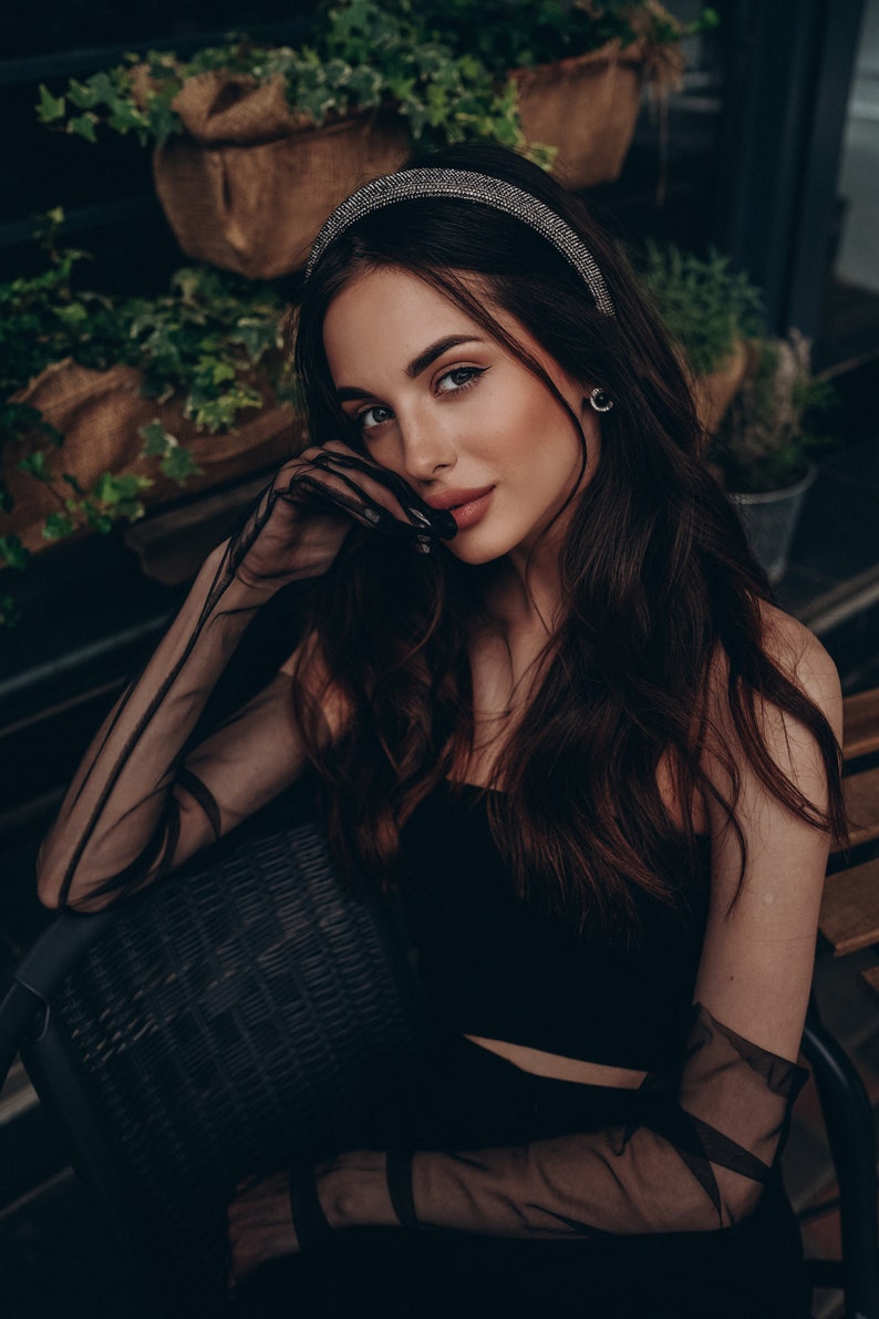 May include: A woman wearing a black top and sheer black gloves with a sparkly headband. She is sitting in a wicker chair.