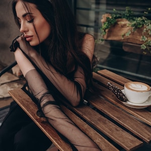 May include: A woman wearing a black sheer top and black pants sits at a wooden table with a cup of coffee and a cookie. She is wearing a black headband with rhinestones.