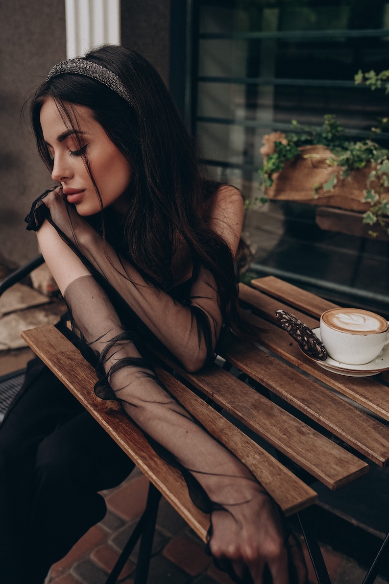 May include: A woman wearing a black sheer top and black pants sits at a wooden table with a cup of coffee and a cookie. She is wearing a black headband with rhinestones.