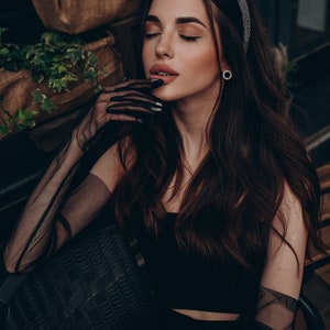 May include: A woman with long dark hair wearing a black top and sheer black gloves. She is sitting in a chair with a wicker back. She is wearing a sparkly headband.
