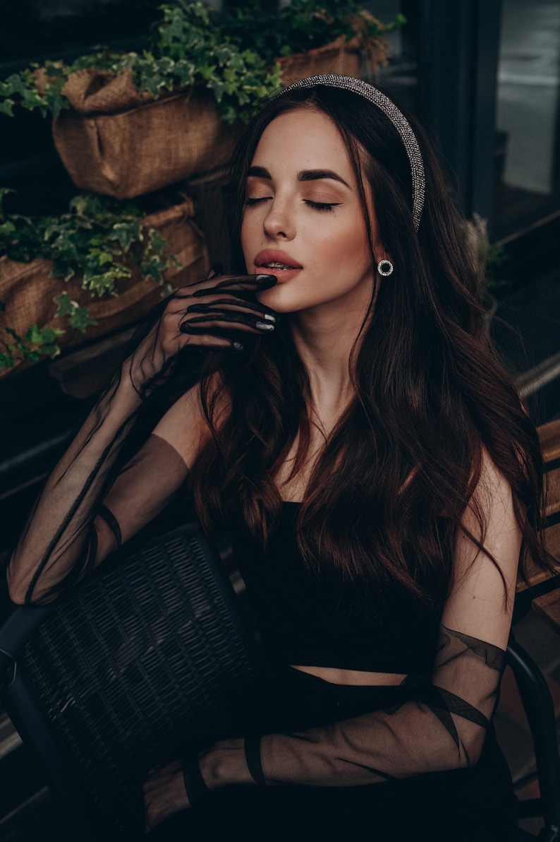 May include: A woman with long dark hair wearing a black top and sheer black gloves. She is sitting in a chair with a wicker back. She is wearing a sparkly headband.
