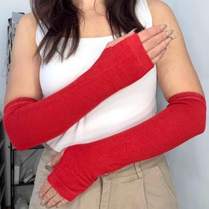 Könnte beinhalten: Ein Paar leuchtend rote Armstulpen aus weichem, fein gestricktem Stoff. Diese langen, fingerlosen Ärmel reichen vom Oberarm bis zu den Knöcheln und verfügen über eine einfache Daumenöffnung sowie ein schlichtes, nahtloses Design.