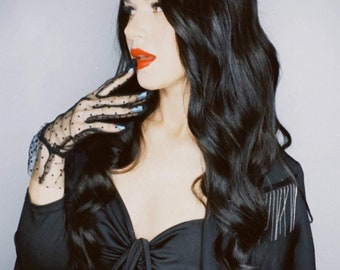 May include: A woman with long, wavy black hair, red lipstick, and blue nail polish. She wears a black top with a bow detail, a black blazer with fringe, and sheer black gloves with polka dots. The image has a vintage aesthetic.
