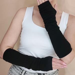 May include: A pair of black arm warmers made of a soft, stretchy fabric. The arm warmers are long enough to cover the entire forearm and hand. They are shown being worn by a person with a white tank top and jeans.