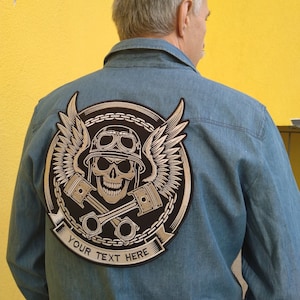 May include: A denim shirt with a large, embroidered patch on the back. The patch features a skull wearing a helmet and goggles, with wings and crossed pistons. The text "YOUR TEXT HERE" is on a banner below the skull.