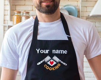 May include: A black apron with the text "Your name" and a graphic of two crossed butcher knives with a red heart and the text "Cooking" in orange.