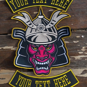 Puede incluir: Un parche bordado negro y amarillo con una cara de demonio roja y rosa que lleva un casco de samurái. El parche tiene el texto "YOUR TEXT HERE" en la parte superior e inferior.