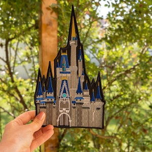 Pode incluir: Um remendo bordado em preto e cinza do icônico Castelo de Cinderela no Walt Disney World. O castelo é detalhado com detalhes azuis e brancos e apresenta uma torre do relógio.