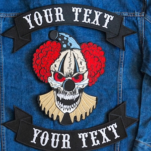 May include: A black denim jacket with two black and white embroidered patches. The patches are banners with the text "YOUR TEXT" in white. The top patch is above a skull with red hair and a blue hat. The skull has a black nose and red eyes.