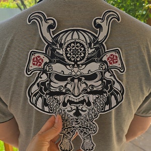 May include: A black and white embroidered patch of a samurai warrior's helmet with a detailed design. The patch is sewn onto a striped gray shirt.