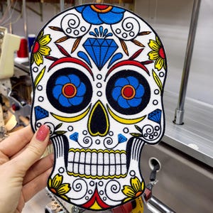 May include: A white skull-shaped patch with colorful embroidery. The design features blue, yellow, red, and black accents, including floral and diamond patterns. The eyes are large blue circles with red centers.