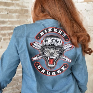 May include: A blue denim shirt with a black and white biker garage patch on the back. The patch features a panther wearing goggles and crossed spark plugs. The text on the patch reads "Bikers Garage".