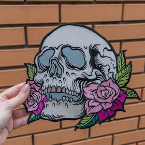 May include: A white skull with a detailed design is surrounded by pink and green roses. The skull has a detailed design with a prominent nose and teeth. The roses are in full bloom and have a realistic look.