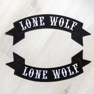 May include: Two black embroidered patches in a banner shape, each with the words "LONE WOLF" in white, stylized lettering. The patches have a textured appearance and are designed to be sewn onto clothing or accessories.