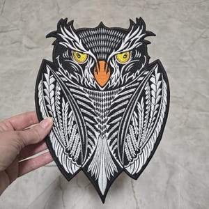 May include: Black and white embroidered owl patch with orange beak and yellow eyes. The owl is surrounded by a white and black patterned design.