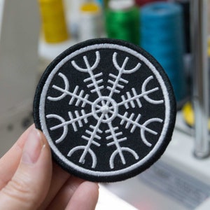 Può includere: Una toppa ricamata in bianco e nero con un design circolare. Il design è una stella bianca a otto punte con un cerchio più piccolo al centro. La toppa ha un diametro di circa 5 cm.