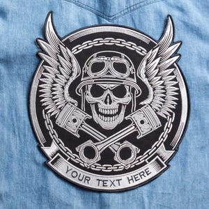 May include: A black and silver embroidered patch featuring a skull wearing a helmet and goggles, with wings and crossed pistons. The patch is surrounded by a chain design and includes the text "YOUR TEXT HERE" on a banner.
