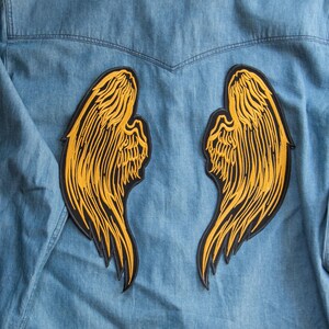 Op de afbeelding: Lichtblauwe spijkerblouse met twee grote, geelzwarte geborduurde engelenvleugelpatches op de rug. De vleugels zijn gedetailleerd met veervormige lijnen en een zwarte rand. De blouse heeft een klassieke kraag en knoopsluiting.