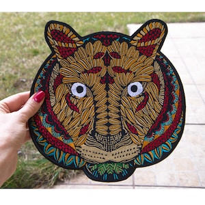 Op de afbeelding: Geborduurde tijgerkop patch met een gedetailleerd ontwerp. De patch heeft een zwarte rand, met een tijgergezicht in tinten goud, rood en teal. De ogen van de tijger zijn wit. Geschikt voor jassen, tassen of andere stoffen items.
