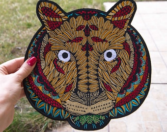 May include: Embroidered tiger head patch with a detailed design. The patch features a black border, with a tiger face in shades of gold, red, and teal. The tiger's eyes are white. Suitable for jackets, bags, or other fabric items.