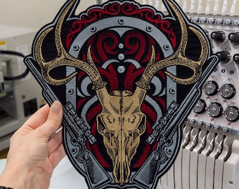 May include: An embroidered patch featuring a tan deer skull with large antlers centred over a red and black decorative shield design. The patch includes two grey rifles positioned diagonally behind the skull and intricate scrollwork.
