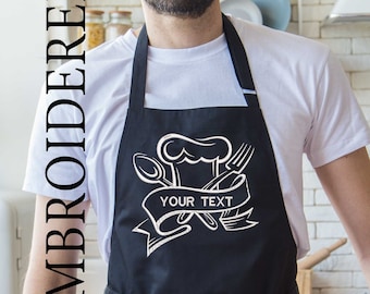 May include: Black apron with a white embroidered design of a chef's hat, spoon, fork, and a banner that says "YOUR TEXT".